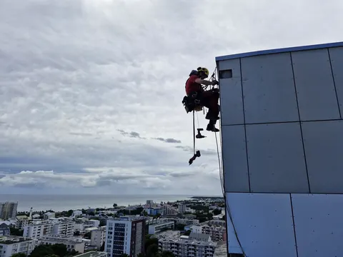 Cordiste suspendu sur ses cordes le long d'une façade en panneaux gris, la ville et la mer en contrebas