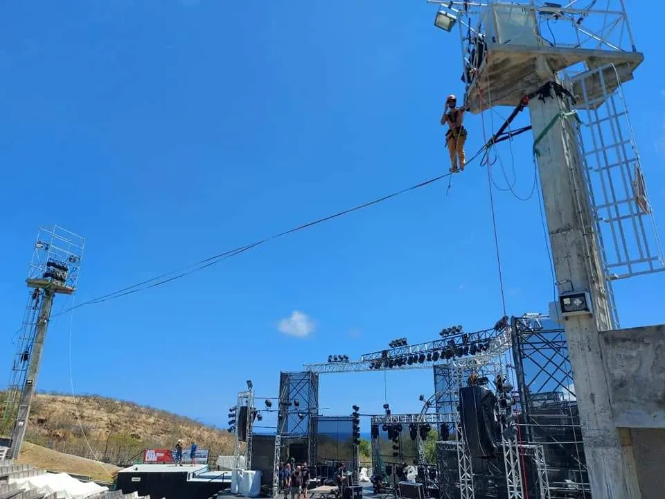 Cordiste debout sur un câble tendu entre deux pylônes d'éclairage, au-dessus de la scène d'un festival en plein air